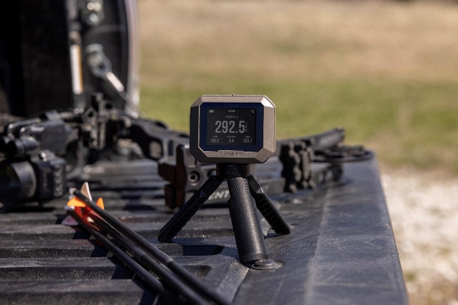 Garmin Xero C1 Pro chronograph on its small tripod, with archery equipment in the background; manufacturer reference photograph.